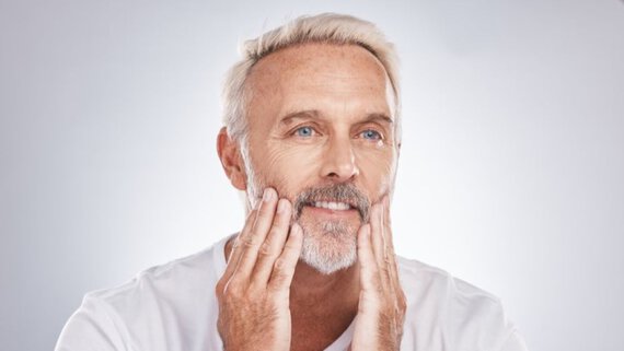 A mature man with white hair and a beard gently touches his face, displaying a thoughtful expression.